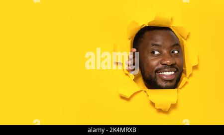 Joyful black man looking aside at copy space, standing in torn yellow paper hole, noticing pleasant unexpected offer Stock Photo