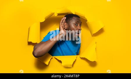 Curious african american man holding hand by his ear, posing through hole in torn yellow background Stock Photo