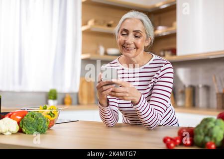 Mobile Communication. Portrait Of Smiling Senior Lady Using Smartphone In Kitchen Interior Stock Photo