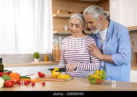 Loving Senior Husband Embracing Wife While She Cooking Food In Kitchen Stock Photo