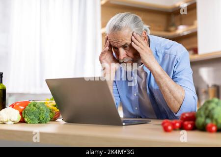 Portrait of stressed senior man using laptop in kitchen interior Stock Photo