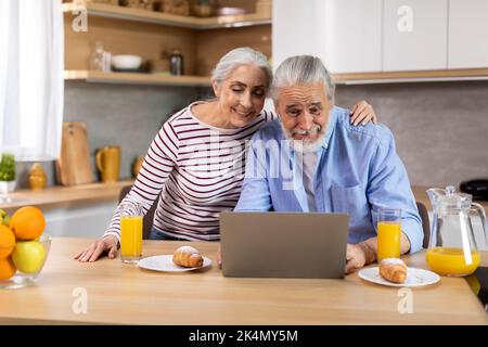 Smiling Elderly Spouses Relaxing With Laptop While Having Breakfast In Kitchen Stock Photo