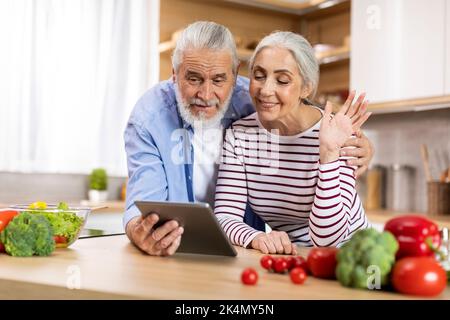 Video Call. Smiling Elderly Couple Using Digital Tablet In Kitchen For Teleconference Stock Photo
