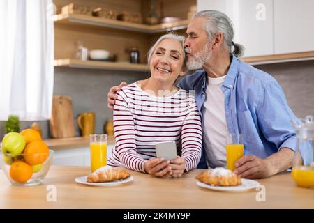 Romantic Happy Senior Couple Resting With Smartphone And Having Breakfast In Kitchen Stock Photo