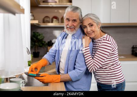 Happy senior couple washing dishes in kitchen while doing cleaning at home Stock Photo