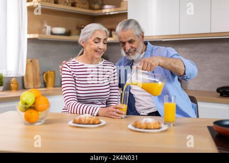 Elderly Spouses Eating Croissants And Drinking Juice While Having Breakfast In Kitchen Stock Photo