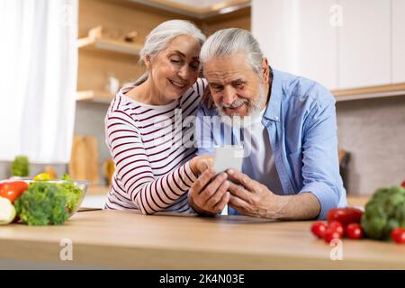 Portrait Of Beautiful Elderly Couple Relaxing With Mobile Phone In Kitchen Interior Stock Photo