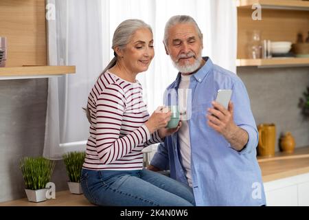 Smiling Elderly Husband And Wife Relaxing In Kitchen With Smartphone And Coffee Stock Photo