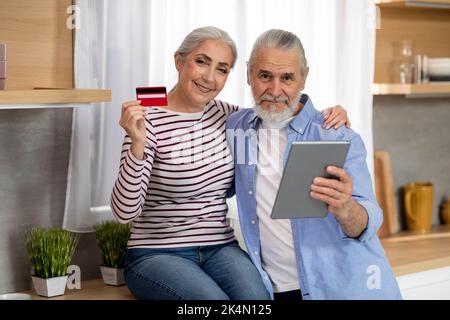 Happy Elderly Couple With Digital Tablet And Credit Card Posing In Kitchen Stock Photo