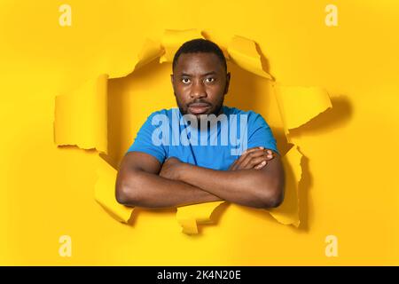 Angry displeased black man feeling outraged keeping arms folded, posing in torn hole of yellow paper background Stock Photo