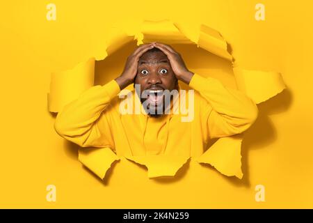 Wow. Portrait of overjoyed black man with open mouth touching head and breaking through hole in yellow paper Stock Photo
