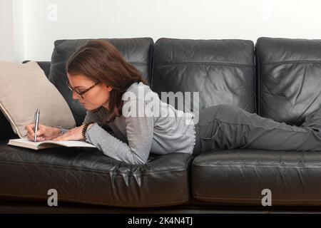 Woman writing in a notebook, laying on sofa. Stock Photo