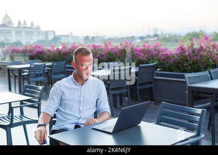 Successful young executive looking while sitting at a table with a laptop on a hotel terrace and finding time for remote control of employees while on Stock Photo