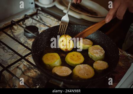 Hands turn over zucchini in flour in frying pan on gas stove Stock Photo