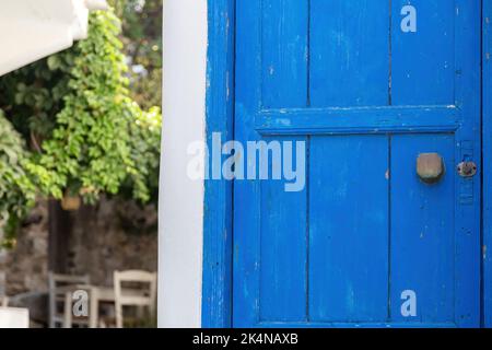 Wooden closed blue old door with rusty handle and keyhole. Greek island vintage entrance, blur traditional cafe, table chairs, plants background. Stock Photo