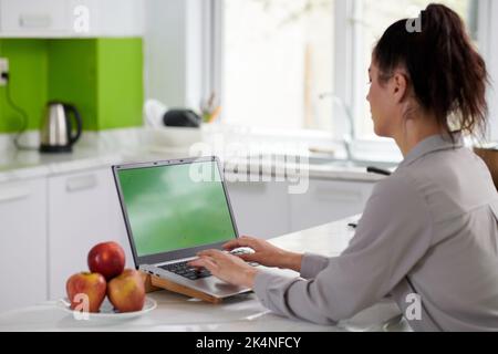 Young modern female creative designer or programmer typing on keypad of laptop with blank green screen while sitting by workplace Stock Photo