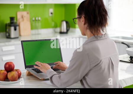 Young brunette female freelancer sitting in front of laptop with blank green screen and typing while using new mobile application Stock Photo