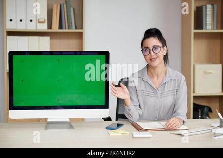 Young brunette businesswoman in eyeglasses and casualwear making presentation of new application while pointing at blank green screen Stock Photo