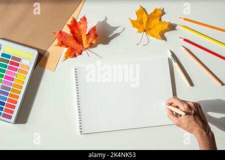 Autumn composition. Woman hand holding brush over blank sketchbook page and begin painting. Watercolor paint, colored pencils and autumn maple fall Stock Photo