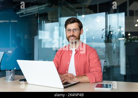 Portrait of mature businessman freelancer startup, bearded man smiling and looking at camera, business owner working inside modern office building wearing red shirt and glasses. Stock Photo