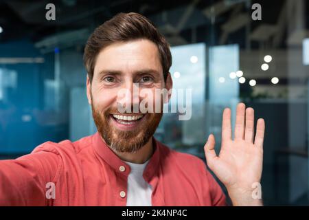 Young happy businessman looking into smartphone camera and talking on video call, man working inside modern office building, smiling and talking remotely with friends, using communication app. Stock Photo