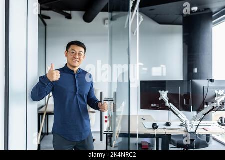 Portrait of Asian designer engineer, man smiling and looking at camera holding thumb up affirmative, businessman working with several computers inside twisted office building. Stock Photo