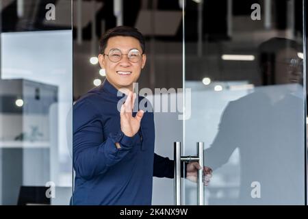 Successful asian worker smiling and looking at camera, man opens door walks into middle of meeting room, holds hand up greets colleagues. Stock Photo