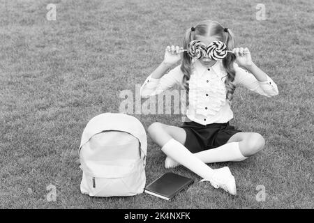 Girl child in school uniform have fun covering eyes with lollipops on green grass, sweet tooth Stock Photo
