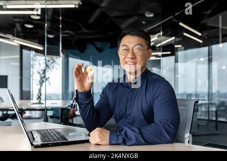 Portrait of satisfied asian broker investor, businessman looking at camera and smiling man holding crypto currency bitcoin virtual money in hands, working inside modern office building. Stock Photo