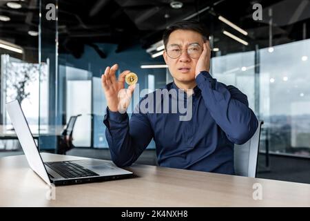Dissatisfied and confused in depression asian financier investor, holding coin money crypto currency bitcoin in hands, male businessman working dissatisfied with exchange rate, broker in despair. Stock Photo