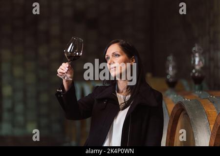 Woman in the wine cellar with barrels in background drinking and tasting wine. Stock Photo