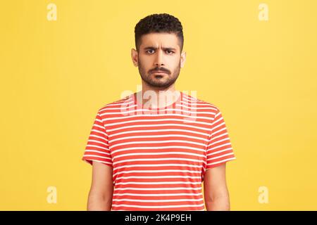 Portrait of serious sad upset man with beard wearing red T-shirt standing looking at camera, expressing sorrow and sadness, hearing bad news. Indoor studio shot isolated on yellow background. Stock Photo