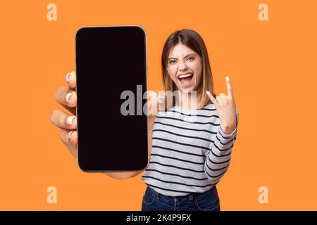 Portrait of excited woman wearing striped shirt standing showing cell phone with empty display, showing rock and roll gesture, screaming happily. Indoor studio shot isolated on orange background. Stock Photo