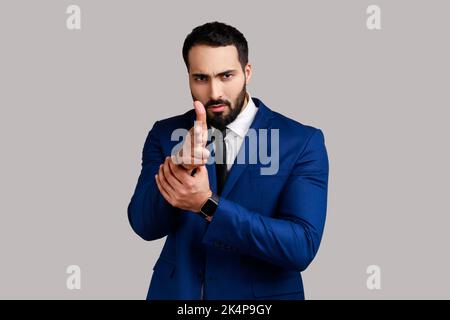 Portrait of serious bearded man imitating guns, shooting into camera with finger pistols, looking menacingly, wearing official style suit. Indoor studio shot isolated on gray background. Stock Photo