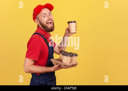 Side view portrait of excited amazed delivery man standing with take away coffee and drinking hot beverage, looking at camera and laughing. Indoor studio shot isolated on yellow background. Stock Photo