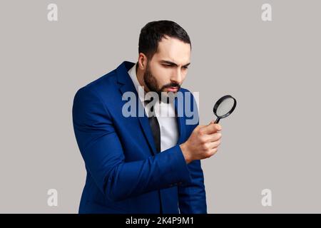 Concentrated curious bearded man looking through magnifying glass, finding out something, exploring, inspecting, wearing official style suit. Indoor studio shot isolated on gray background. Stock Photo