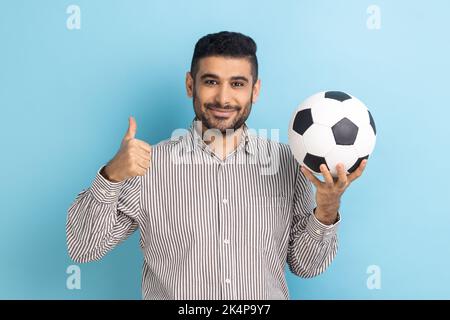 Satisfied bearded businessman holding soccer ball on his hand with smiling positive expression, showing thumb up, wearing striped shirt. Indoor studio shot isolated on blue background. Stock Photo