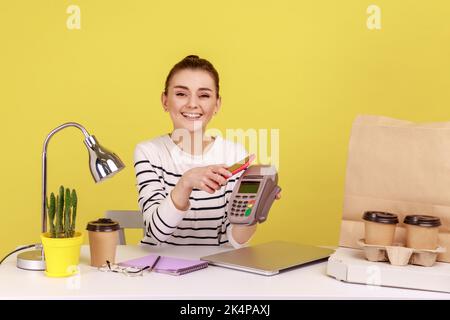 Happy young woman sitting at workplace with mobile smart phone and paying by pay pass pos. contactless, nfc technology on cellphone and payment. Indoor studio studio shot isolated on yellow background Stock Photo