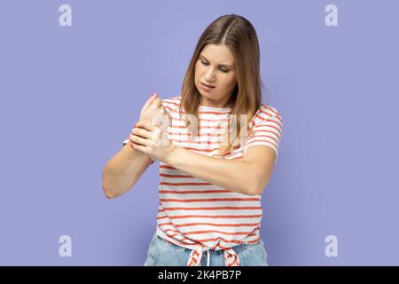 Unhappy sick blond woman wearing striped T-shirt touching painful hand, suffering trauma, sprain wrist, feeling ache of carpal tunnel syndrome. Indoor studio shot isolated on purple background. Stock Photo