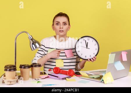 Serious shocked woman office worker pointing finger at big wall clock in her hands sitting at workplace, working late hours, overtime job. Indoor studio studio shot isolated on yellow background. Stock Photo