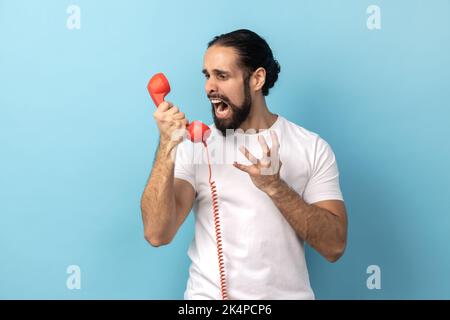 Portrait of angry man with beard wearing white T-shirt screaming and yelling talking retro landline phone, complaining on connection quality. Indoor studio shot isolated on blue background. Stock Photo