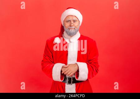 Portrait of elderly man with gray beard wearing santa claus costume standing with folded hands and looking at camera with upset expression. Indoor studio shot isolated on red background. Stock Photo