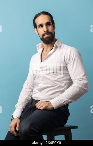 Unsmiling serious dark haired attractive man with beard wearing white shirt and black trousers, looking away with attentive look while sitting on chair. Indoor studio shot isolated on blue background. Stock Photo