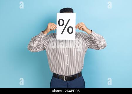 Portrait of unknown anonymous businessman hiding his face behind paper with percent sign inscription, wearing striped shirt. Indoor studio shot isolated on blue background. Stock Photo