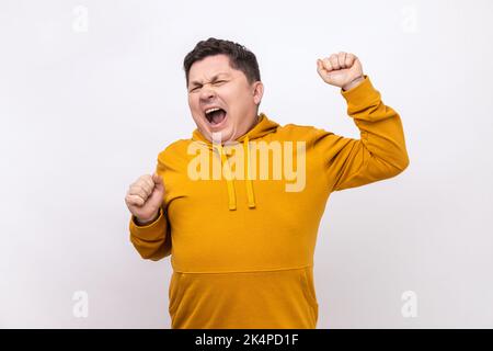 Portrait of middle aged attractive man yawning and raising hands up, feeling fatigued of overtime work, wearing urban style hoodie. Indoor studio shot isolated on white background. Stock Photo