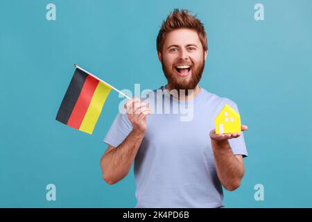Portrait of positive optimistic handsome bearded man holding toy paper house and flag of germany in hand, moving into another country. Indoor studio shot isolated on blue background. Stock Photo