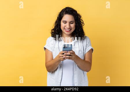 Portrait of smiling satisfied young adult woman with dark wavy hair using mobile phone with happy expression, checking social networks. Indoor studio shot isolated on yellow background. Stock Photo