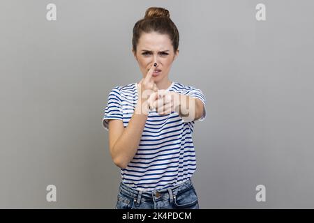 Hey, you are liar. Portrait of woman wearing striped T-shirt standing with upset disappointed face and touching nose, showing lie gesture. Indoor studio shot isolated on gray background. Stock Photo