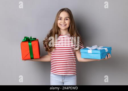 Portrait of winsome adorable little girl wearing striped T-shirt celebrating her birthday, holding two present boxes in hands and looking at camera. Indoor studio shot isolated on gray background. Stock Photo