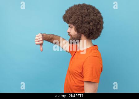 Side view of displeased man with Afro hairstyle wearing orange T-shirt showing thumbs down dislike gesture, symbol of disagree, giving feedback. Indoor studio shot isolated on blue background. Stock Photo